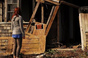 Young woman hesitates outside of dilapidated old farmhouse with a KEEP OUT sign