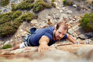 man reaching for a grip while he rock climbs on a steep cliff